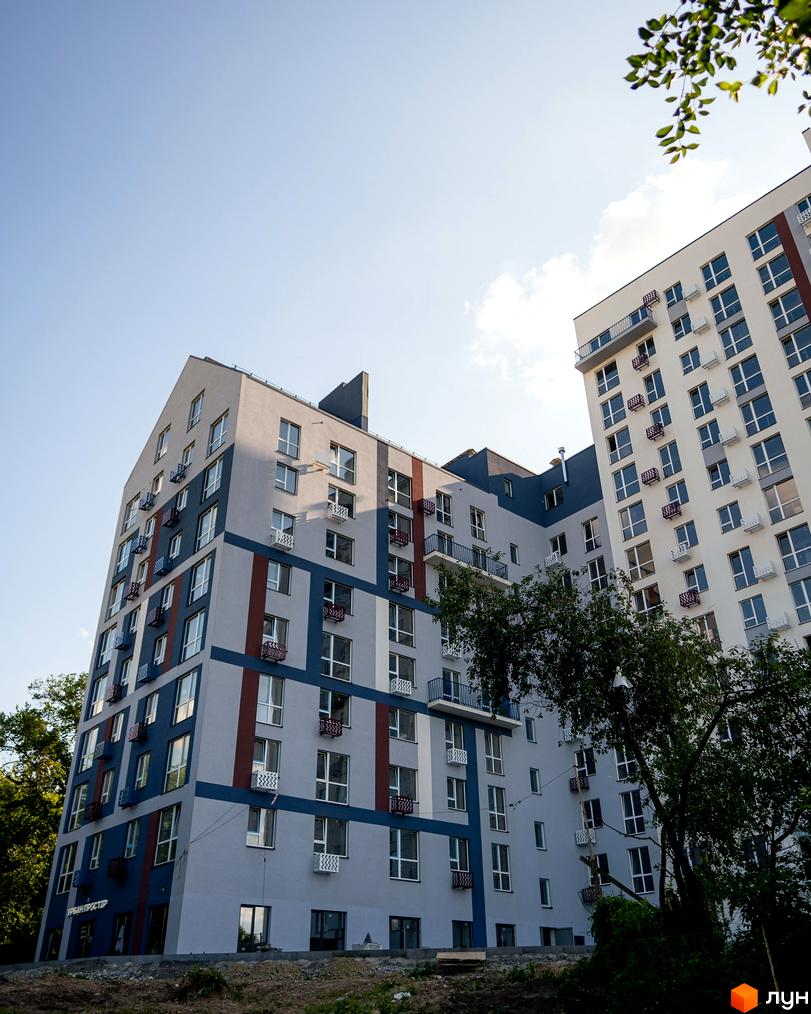 Новобудова ЗиґЗаґ: багатоповерхові будинки зі світлими фасадами, синіми та червоними смугами, численними вікнами та балконами. Поруч ростуть дерева.