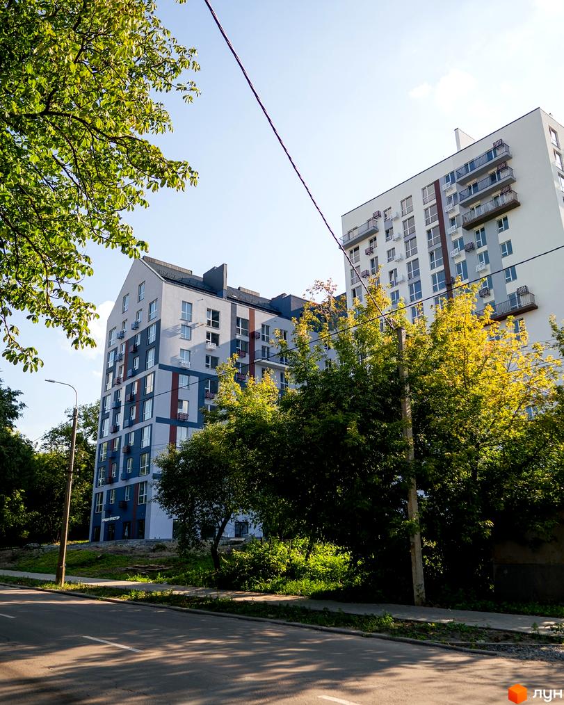 Житловий комплекс ЗиґЗаґ: багатоповерхові будинки світло-сірого та білого кольору з балконами, оточені зеленими деревами, дорогою та тротуаром.