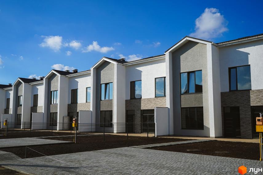 Таунхауси Lazur Village з білими та сірими фасадами, великими вікнами та брукованою прибудинковою територією під блакитним небом.