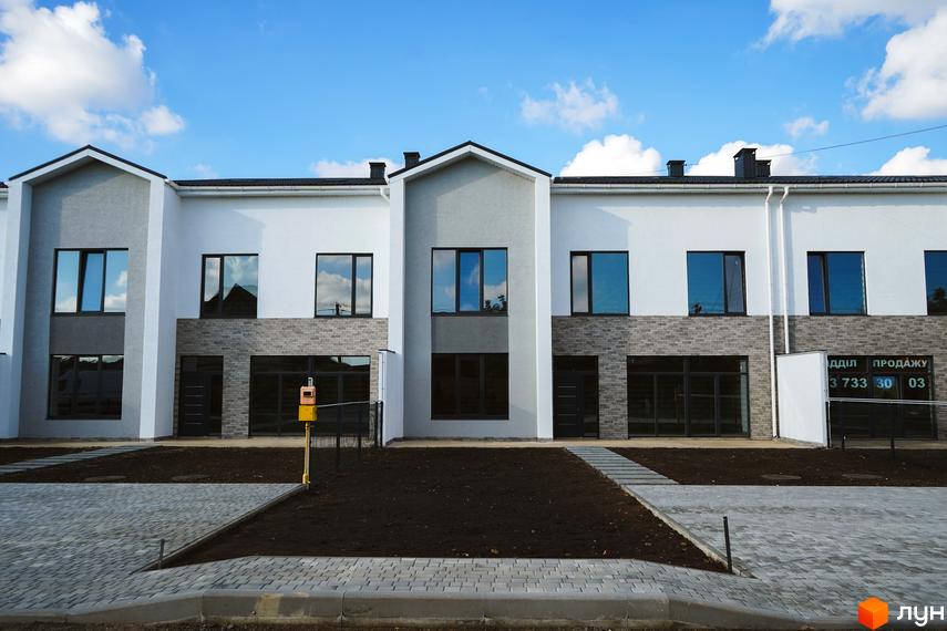 Таунхауси Lazur Village з сірою та білою штукатуркою, цегляним оздобленням та великими вікнами. Перед будинками облаштована прибудинкова територія з бруківкою та газоном.