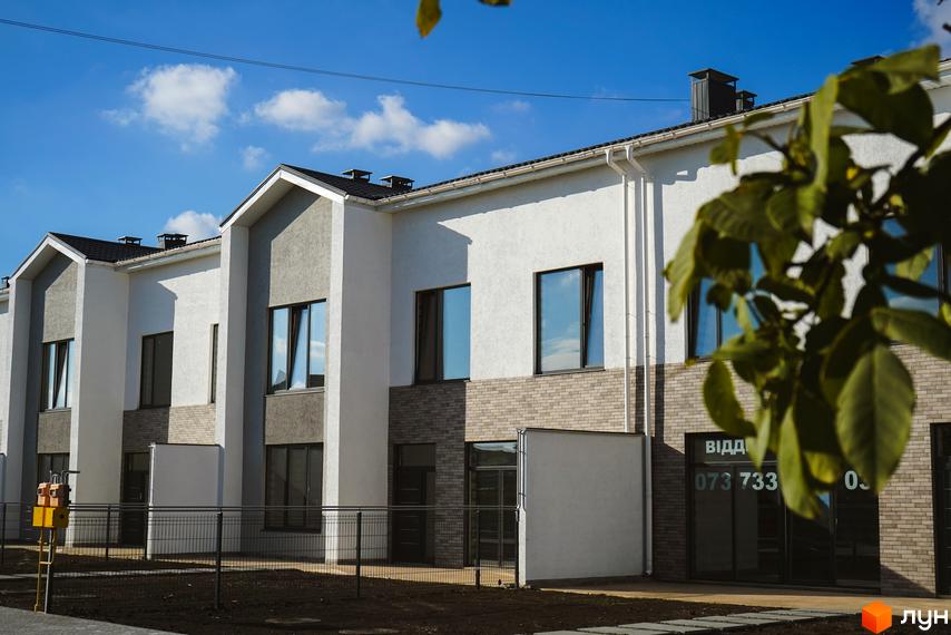 Зовнішній вигляд таунхаусів Lazur Village з білими фасадами та сірими акцентами, вікнами та цегляною кладкою. Прибудинкова територія з огорожею та зеленими насадженнями.