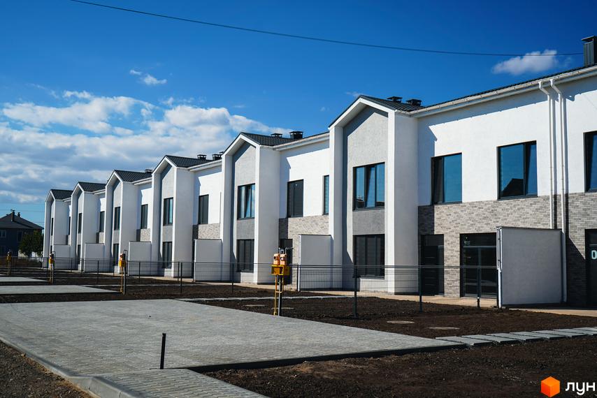 Ряд двоповерхових таунхаусів Lazur Village з білими фасадами та сірими акцентами, вікнами та цегляною обробкою. Поруч облаштована прибудинкова територія з бруківкою та огорожею.