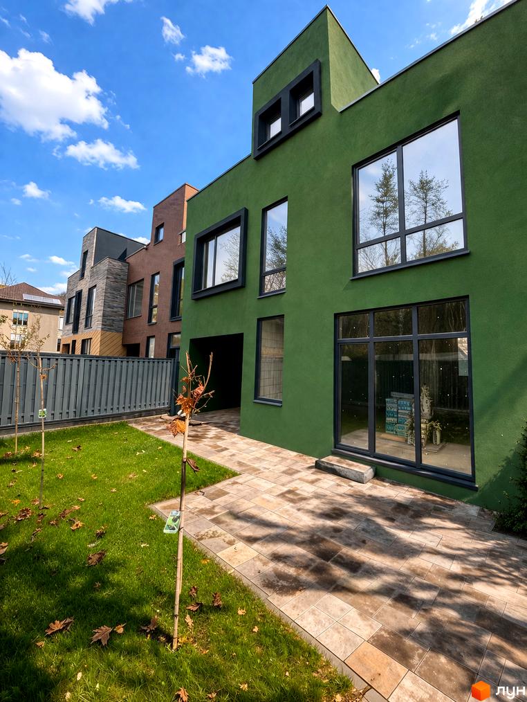 Таунхауси Madera House з зеленою фасадною штукатуркою та великими вікнами, перед якими розташований доглянутий газон та вимощена плиткою доріжка.