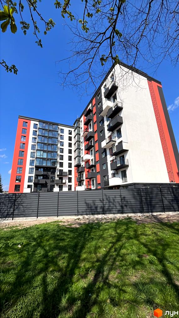 Новобудова вул. Степана Бандери, 60 з червоно-чорно-білим фасадом та балконами. Навколо зелена трава та паркан.