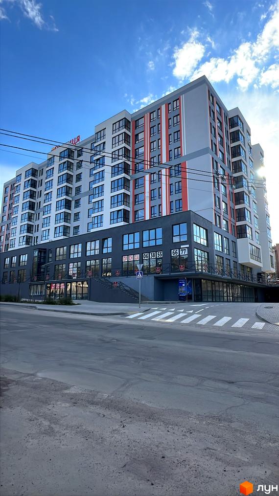 Зовнішній вигляд новобудови на вул. Привокзальна, 30, з фасадом сірого, білого та червоного кольорів, великими вікнами та комерційними приміщеннями на першому поверсі. Поруч розташована дорога з пішохідним переходом.