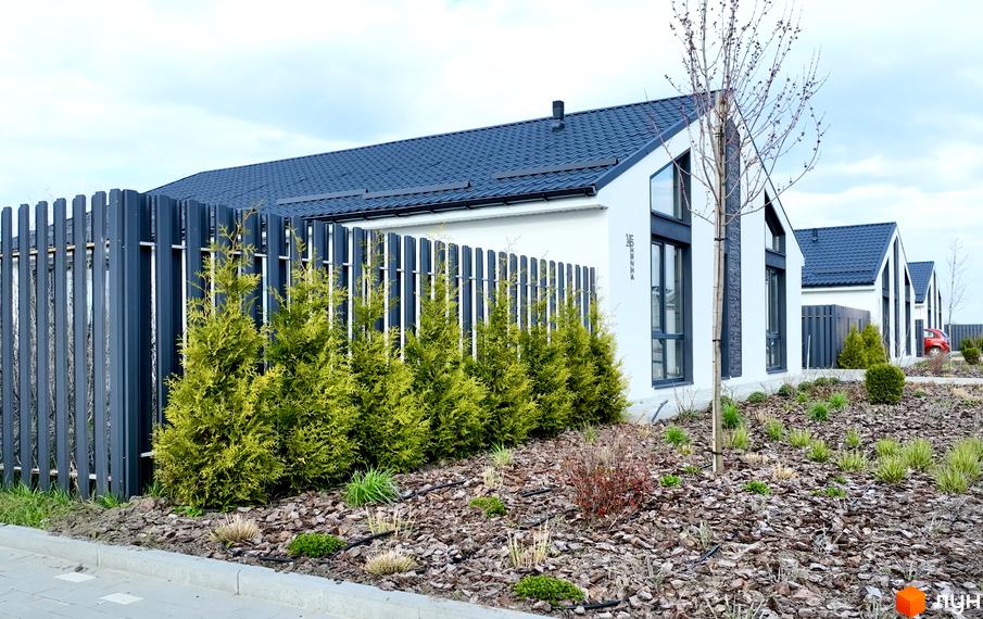 Котеджне містечко КМ Gardd House Eco Village. Зовнішній вигляд будинків з темним дахом та білими стінами, огороджених парканом, та доглянута прибудинкова територія з туями та декоративними рослинами.