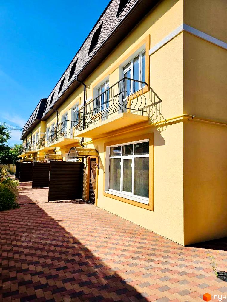 Зовнішній вигляд жовтих таунхаусів Родинний Преміум 2 з балконами та коричневими воротами. Прибудинкова територія вимощена бруківкою.