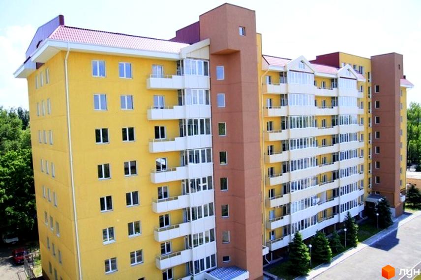 Житловий комплекс Скеля – це яскрава жовта багатоповерхівка з коричневими акцентами та білими балконами. Навколо будівлі розташовані зелені дерева та доглянута прибудинкова територія.