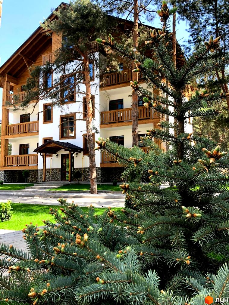 Житловий комплекс DESNA residence з дерев'яними балконами та білими фасадами, оточений соснами. Навколо будинку зелена галявина та доріжки.