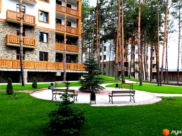 Житловий комплекс DESNA residence з кам'яним фасадом та дерев'яними балконами, оточений соснами та доглянутою зеленою територією з лавками.