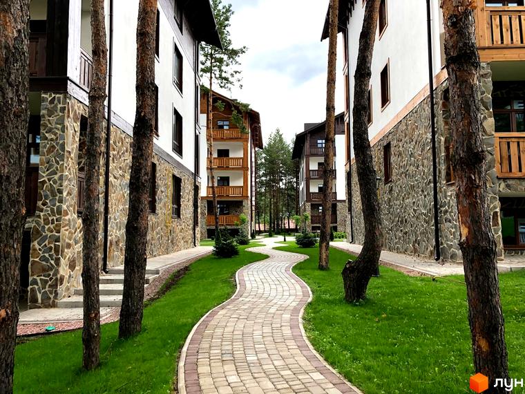 Житловий комплекс DESNA residence з кам'яним фасадом та дерев'яними балконами, оточений зеленою галявиною та доріжкою з бруківки.