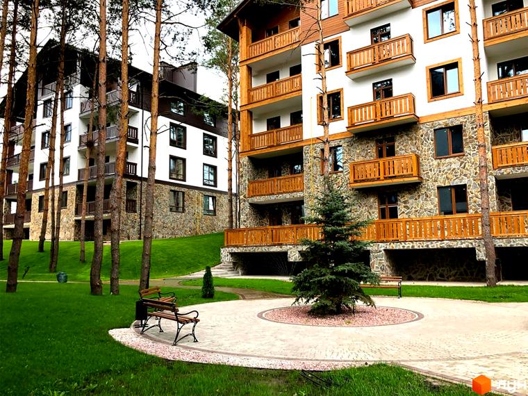 Житловий комплекс DESNA residence з дерев'яними балконами та кам'яним оздобленням фасаду, оточений соснами та доглянутою зеленою територією з лавками.