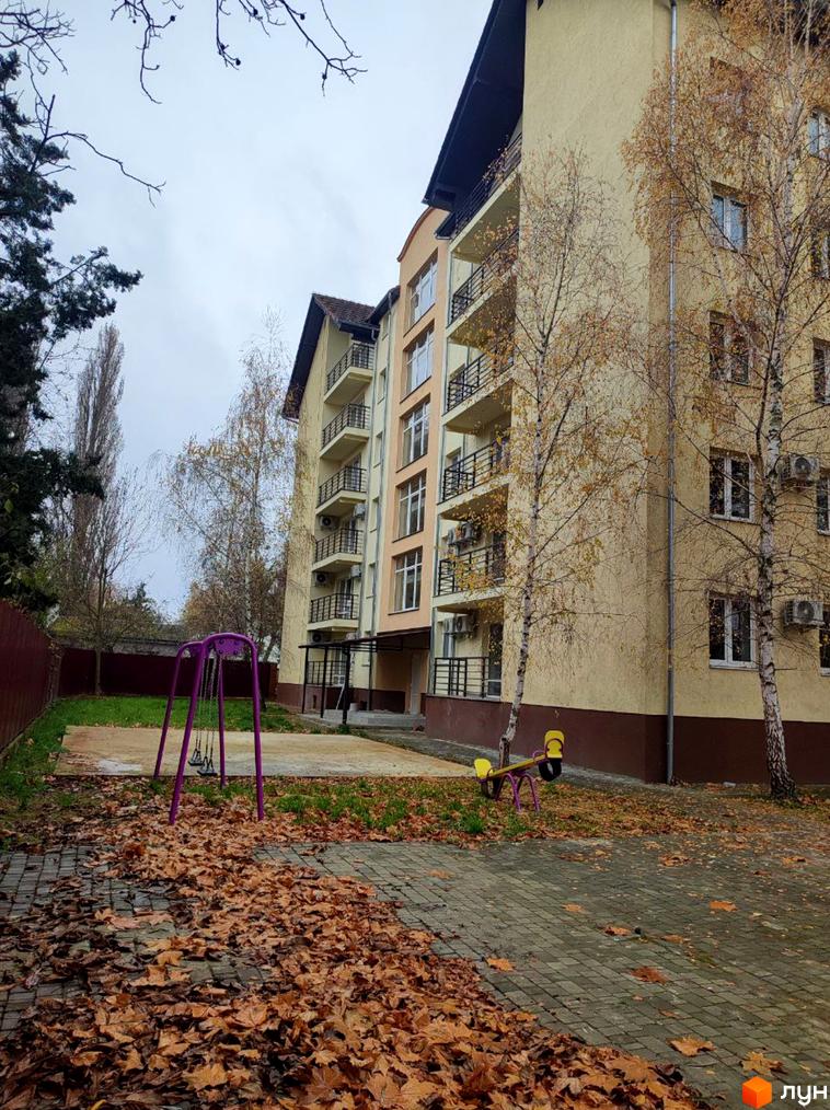 Багатоквартирний будинок світло-жовтого кольору з коричневою основою та балконами. На прибудинковій території дитячий майданчик з гойдалками та опале листя.