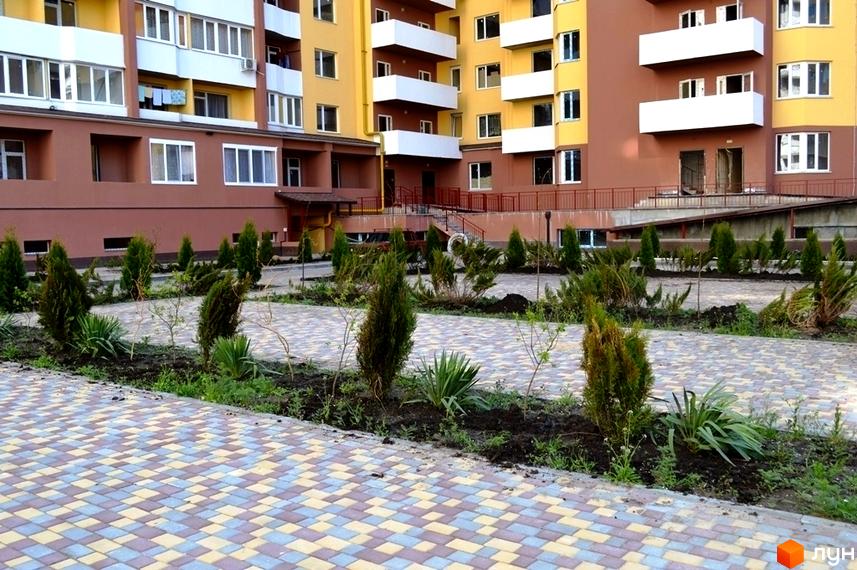 Зображення житлового комплексу з фасадом у коричнево-жовтих тонах. На території перед будинком висаджені зелені насадження та викладена бруківка.