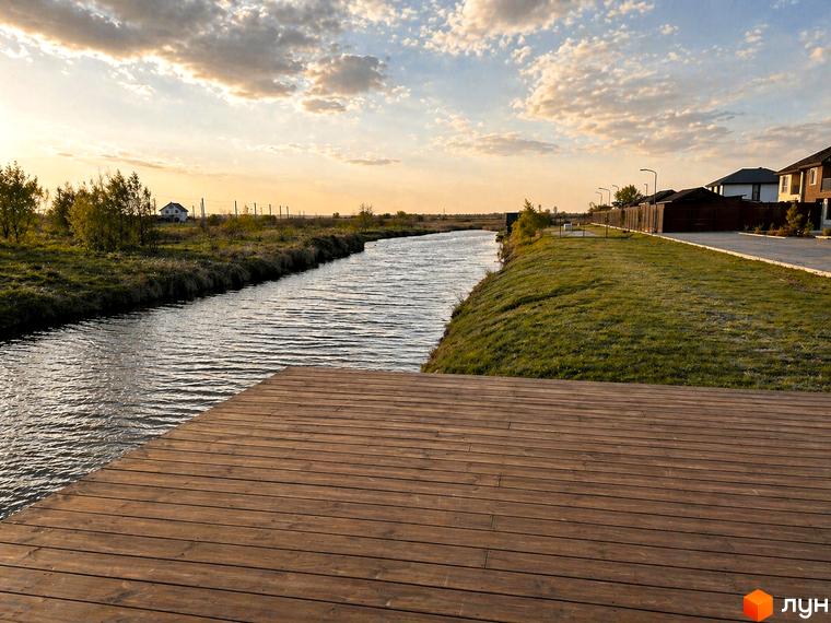 Котеджне містечко DESNA HOMES: котеджі зі світлими фасадами та темними дахами вздовж річки. Територія має зелені газони та дерев'яний настил біля води.
