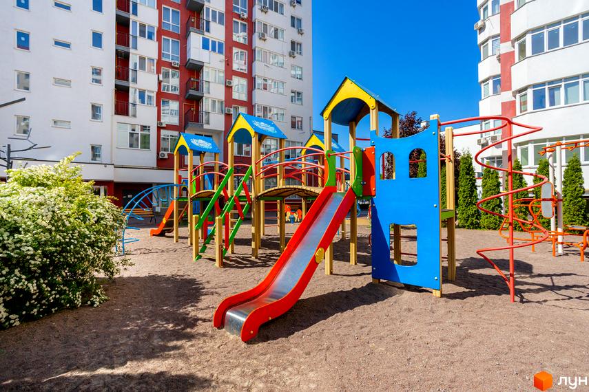 Житловий комплекс Сакура з багатоповерховими будинками білого та червоного кольорів. На передньому плані яскравий дитячий майданчик з гірками та драбинами.