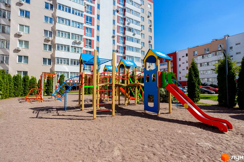 Житловий комплекс Сакура: дитячий майданчик біля багатоповерхових будинків. Зелені насадження та піщане покриття на території.