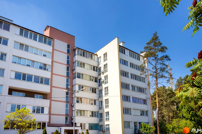 Житловий комплекс Українка - багатоповерховий будинок з білими та рожевими фасадами, вікнами та балконами. Навколо зелені дерева та блакитне небо.