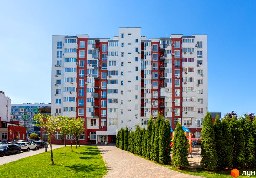 Зовнішній вигляд новобудови ЖК Сакура з червоно-білим фасадом та балконами. Прибудинкова територія з зеленим газоном, деревами та дитячим майданчиком.
