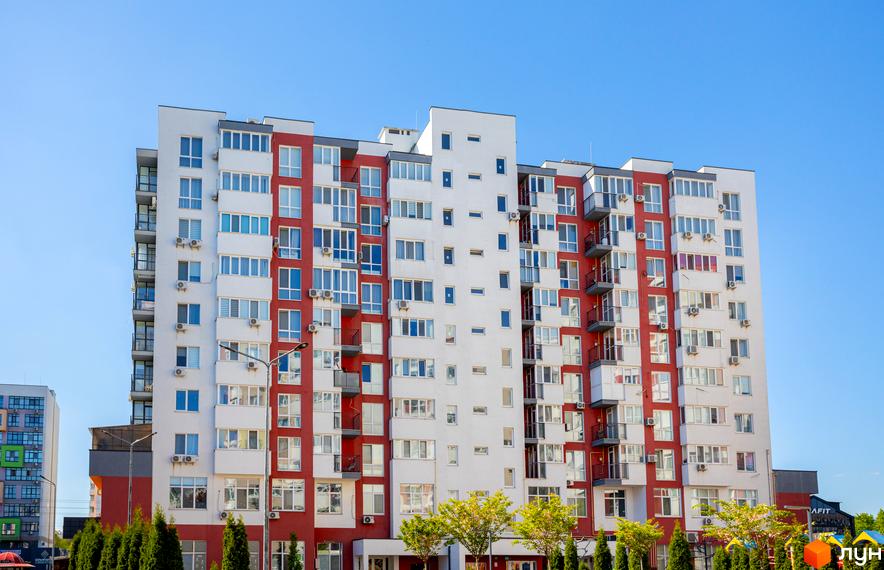 Фасад Житлового комплексу Сакура з білими та червоними вертикальними секціями, численними вікнами та балконами. На передньому плані видно зелені насадження.