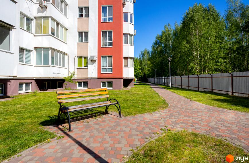 Фасад Новобудови Сакура має білі, бежеві та червоні секції з вікнами. Прибудинкова територія з газоном, лавкою та брукованою доріжкою веде до лісу.