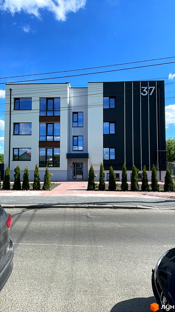 Житловий будинок за адресою вул. Поповича, 37, з білим та темно-сірим фасадом, великими вікнами. На фасаді видно номер 37, а перед будинком – бруківка та декоративні дерева.