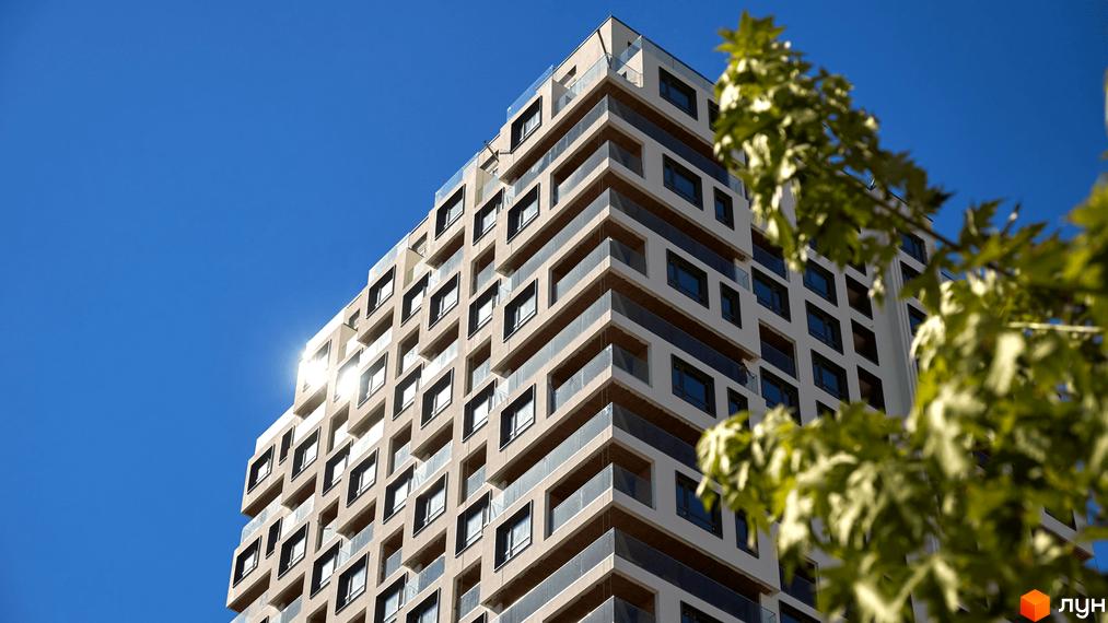 Багатоповерховий будинок Kub29 має світлий фасад з темними вікнами та скляними балконами. На передньому плані видно зелене дерево на фоні синього неба.