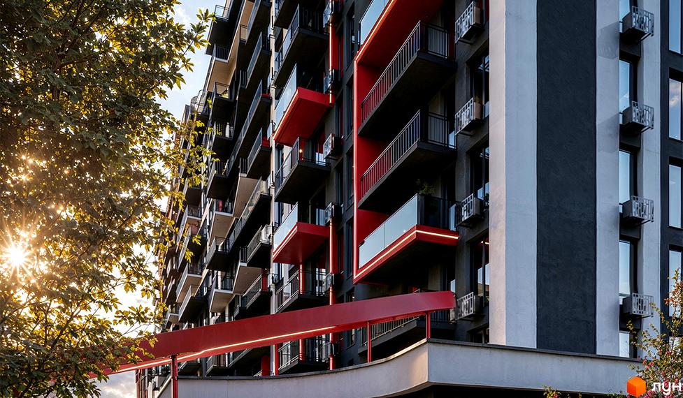 Фасад житлового комплексу Family Plaza з червоними елементами та скляними балконами, освітлений сонцем. Навколо зелені дерева.