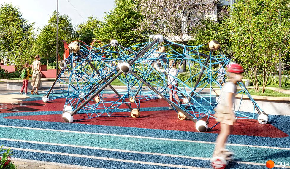 Дитячий майданчик з мотузковим атракціоном у ЖК Comfort Park 2. Територія біля будинку з зеленими деревами та квітучими кущами.