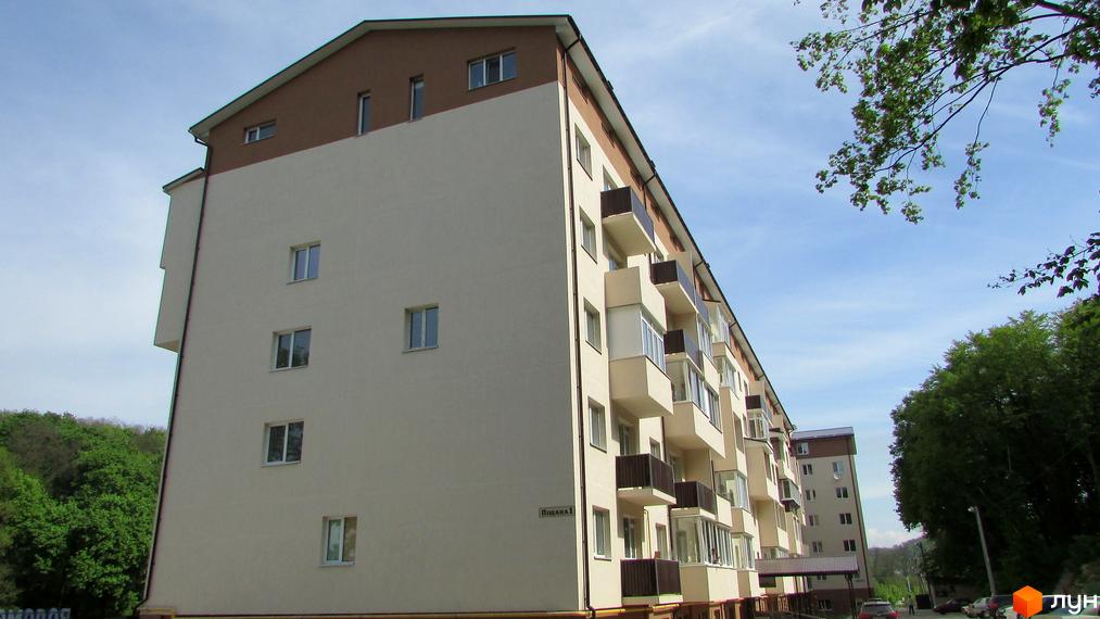 Зовнішній вигляд новобудови на вул. Піщана, 1. Фасад будинку світло-бежевий з коричневим дахом, багато балконів. Навколо зелені дерева.