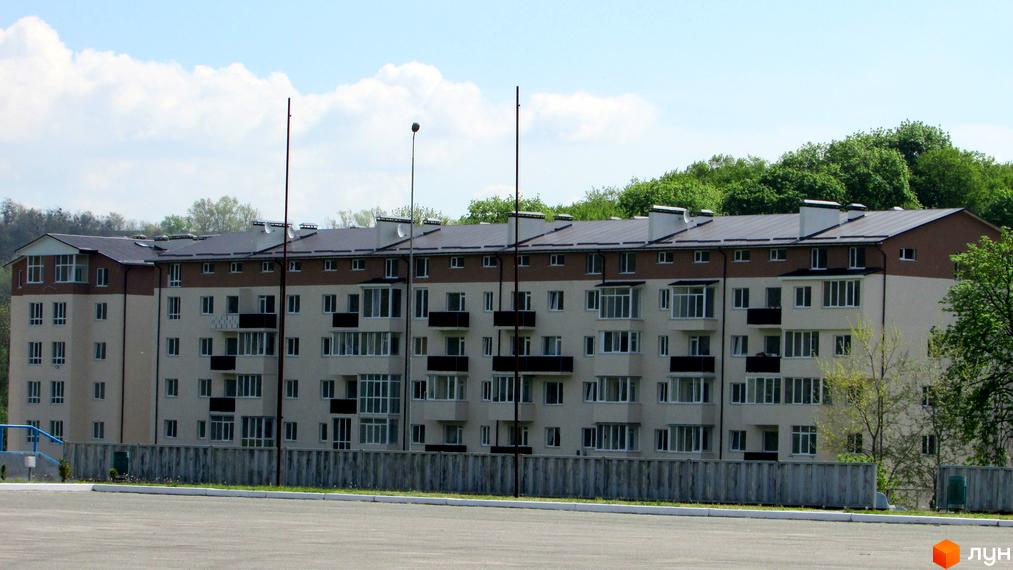 Зовнішній вигляд новобудови на вулиці Піщаній, 1. Багатоповерховий будинок з балконами та вікнами на тлі зелених дерев та блакитного неба.