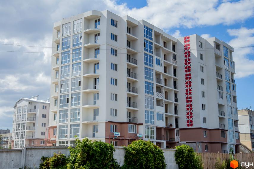 Новобудова ЖК Вишиванка з панорамними вікнами та балконами. На фасаді будівлі присутній орнамент у вигляді вишиванки. Навколо будинку зелені насадження та паркан.