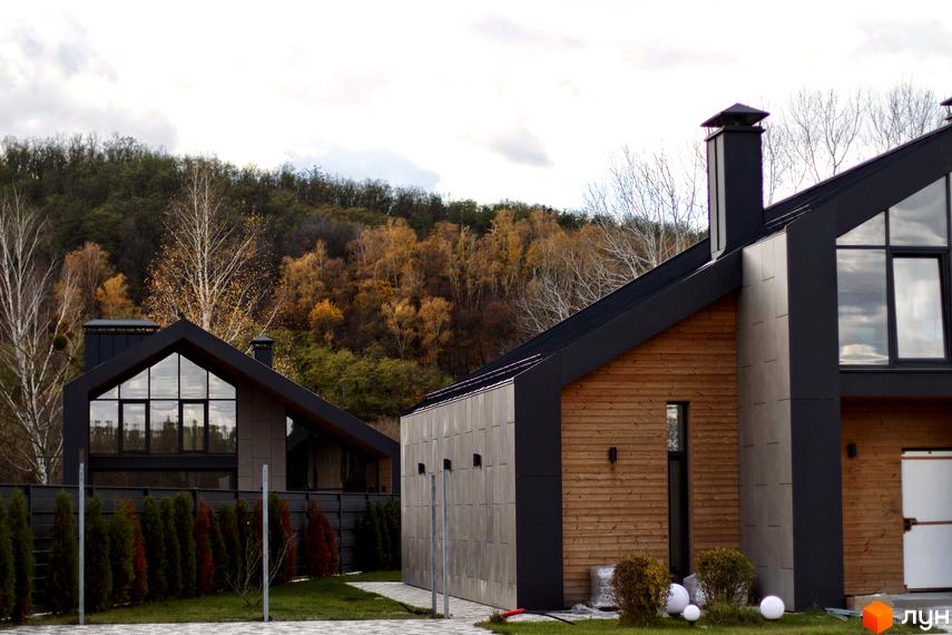 Котеджне містечко КМ Piemont eco village з дерев'яним фасадом та великими вікнами на тлі осіннього лісу. Прибудинкова територія з парканом та зеленими насадженнями.