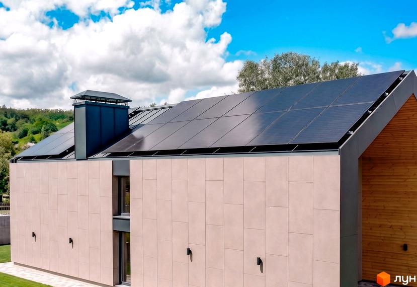 Котеджне містечко КМ Piemont eco village з сонячними панелями на даху та фасадом з світлої плитки. Навколо зелені дерева та блакитне небо з хмарами.