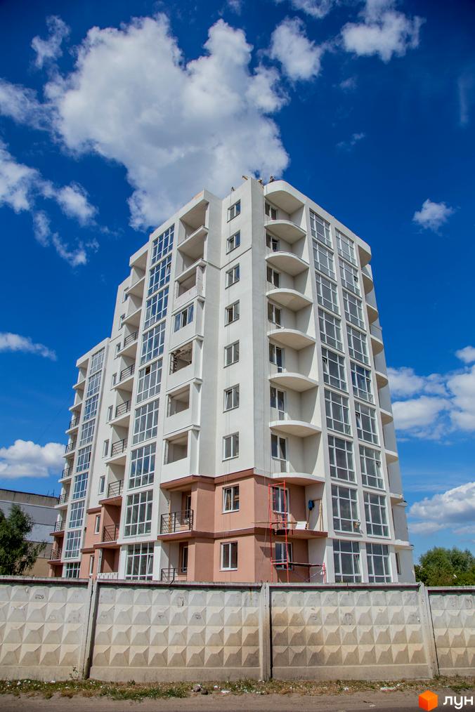 Житловий комплекс Вишиванка. Сучасна багатоповерхова новобудова з панорамними вікнами та балконами на фоні блакитного неба з хмарами. Перед будинком бетонний паркан.