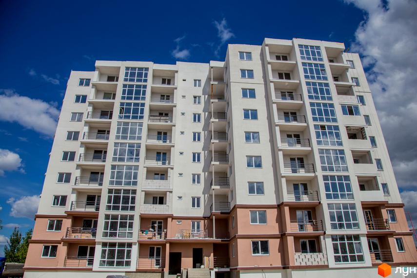 Фасад новобудови ЖК Вишиванка з великою кількістю вікон та балконів. Будинок має білий та рожевий кольори, розташований на фоні блакитного неба з хмарами.