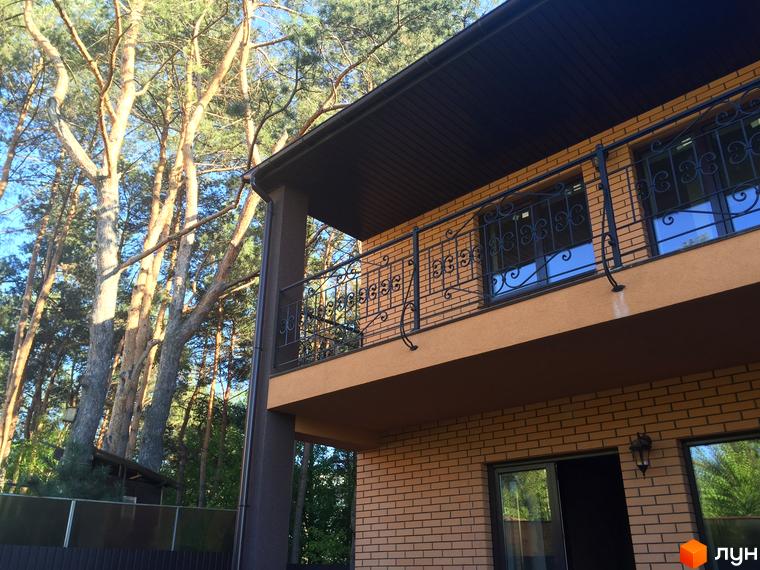 Зовнішній вигляд двоповерхового будинку з цегляним фасадом та балконами з кованими перилами. Навколо будинку високі дерева.