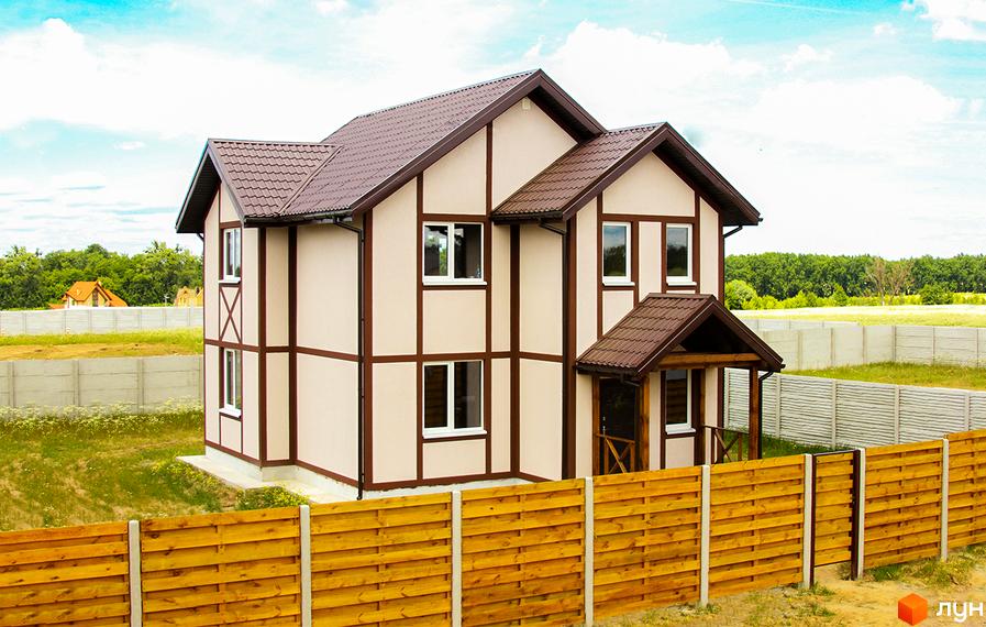Котеджне містечко КМ Альпійський Хуторок: двоповерховий будинок світло-бежевого кольору з коричневими декоративними елементами та коричневим дахом. Навколо будинку зелена трава та дерев'яна огорожа.