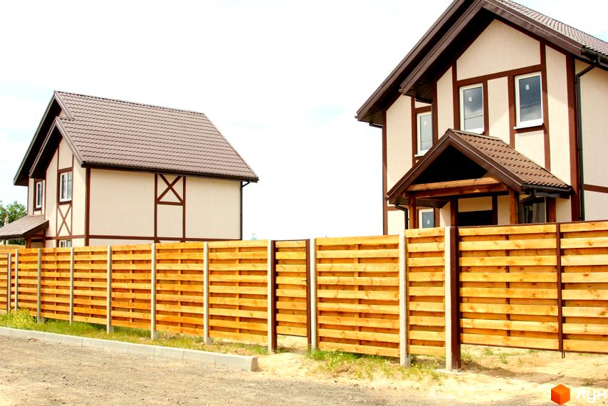 Котеджне містечко КМ Альпійський Хуторок: двоповерхові будинки з коричневими дахами та світлими фасадами, огороджені дерев'яним парканом. Прибудинкова територія з газоном та дорогою.