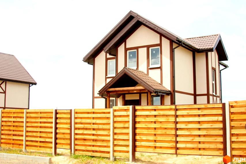 Котеджне містечко КМ Альпійський Хуторок: двоповерхові будинки з коричневими дахами та оздобленням, огороджені дерев'яним парканом. Навколо будівель зелена трава та біле небо.