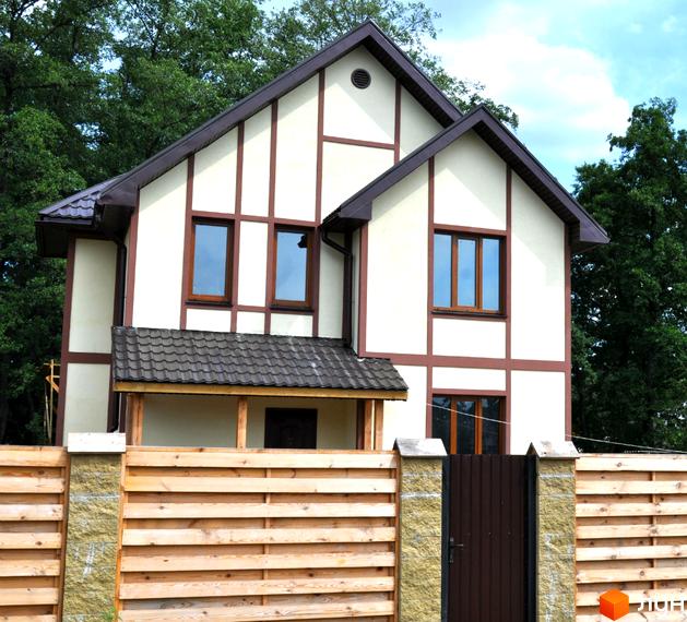 Котеджне містечко КМ Альпійський Хуторок. Двоповерховий будинок з коричневою черепицею та світлим фасадом, огороджений дерев'яним парканом.