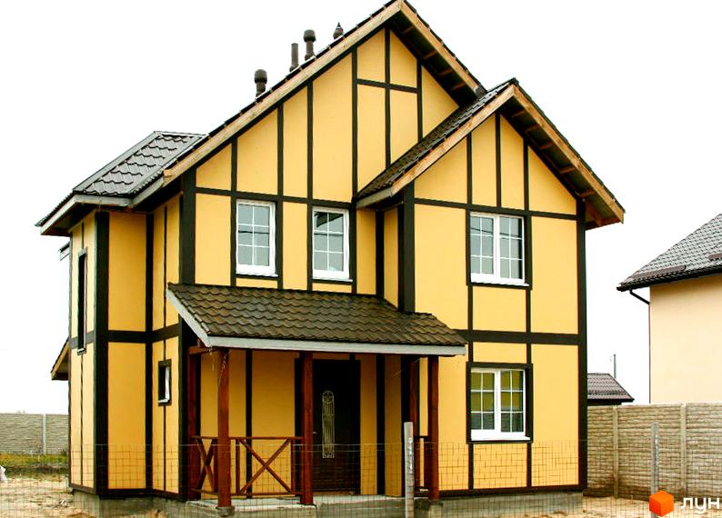 Котеджне містечко КМ Альпійський Хуторок. Двоповерховий будинок жовтого кольору з коричневими балками та темним дахом, вікна з білими рамами, крите крильце біля входу.