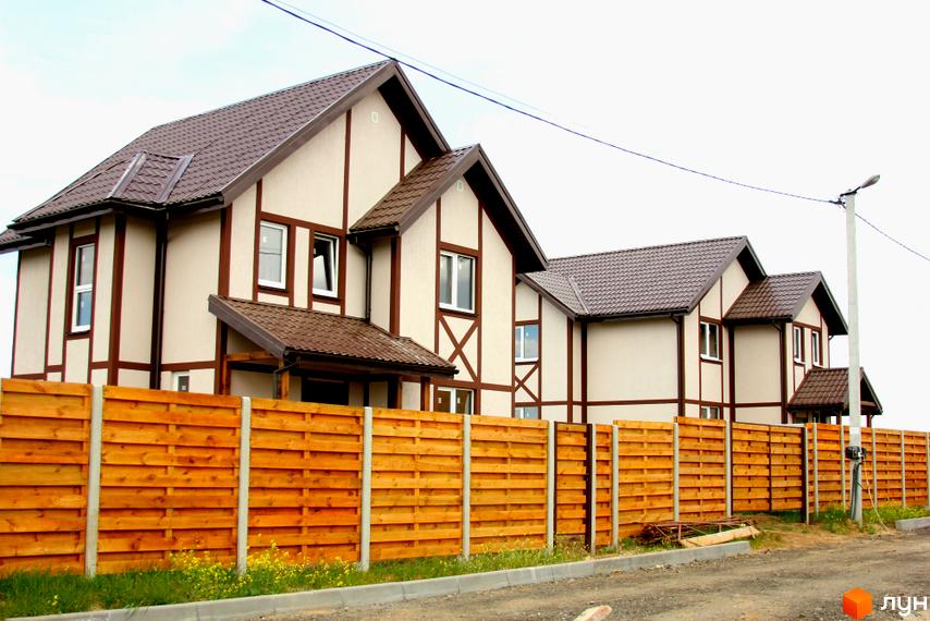 Котеджне містечко КМ Альпійський Хуторок. Двоповерхові будинки з коричневим дахом та світлими стінами, огороджені дерев'яним парканом. Навколо зелена трава та жовті квіти.