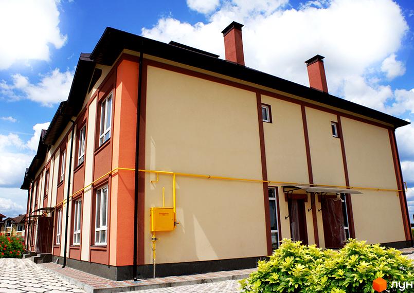 Таунхаус СанЛенд. Фасад двоповерхового будинку з червоною та бежевою обробкою, вікнами та газовою трубою. Поруч доглянутий кущ та бруківка.