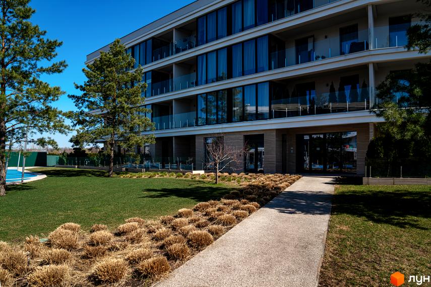 Багатоповерхова будівля Житлового комплексу Park Lake City з великими вікнами та скляними балконами. Перед будинком зелений газон, доріжка та хвойні дерева.