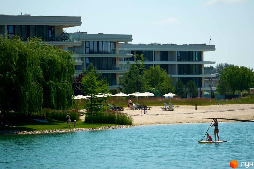 Житловий комплекс Park Lake City з багатоповерховими будинками та пляжною зоною біля озера. Комплекс має сучасну архітектуру та доглянуту прибудинкову територію з зеленню.