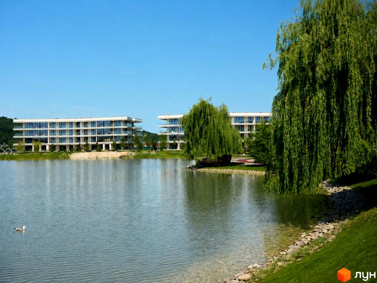 Житловий комплекс Park Lake City з сучасними будівлями та озером. Навколо комплексу розташований доглянутий ландшафт з деревами та зеленою травою.