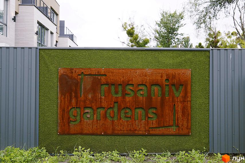 Зовнішній вигляд таунхаусів Rusaniv Gardens з табличкою комплексу на фоні зеленої стіни та дерев. Прибудинкова територія з травою та рослинами.