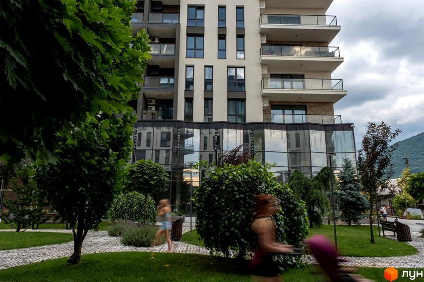 Будинок житлового комплексу SKY CITY з балконами та скляним фасадом. Навколо зелена прибудинкова територія з деревами та доріжками.