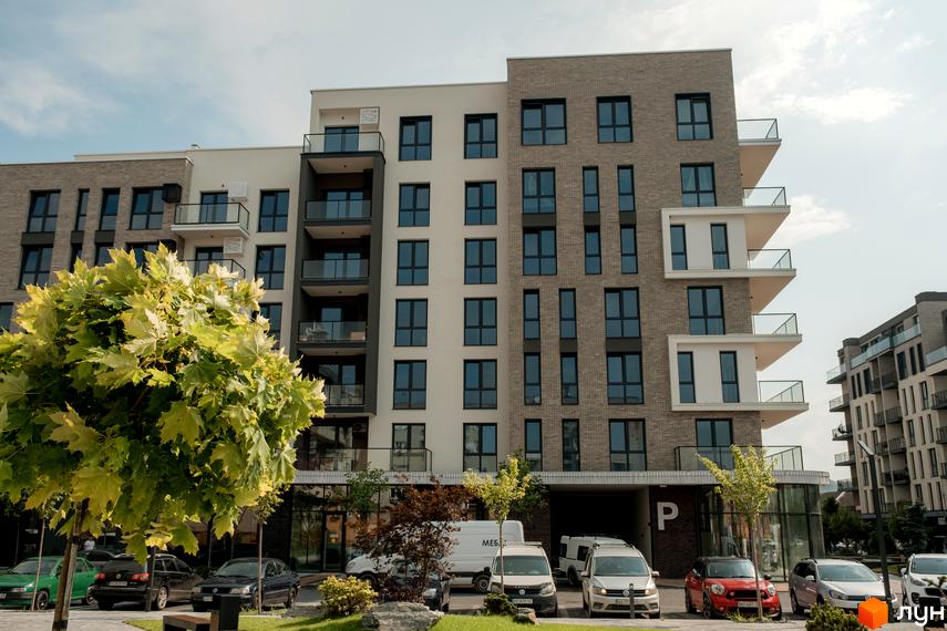 Житловий комплекс SKY CITY: багатоповерховий будинок з комбінованим фасадом, темними вікнами та скляними балконами. На прибудинковій території зелені дерева та парковка з автомобілями.